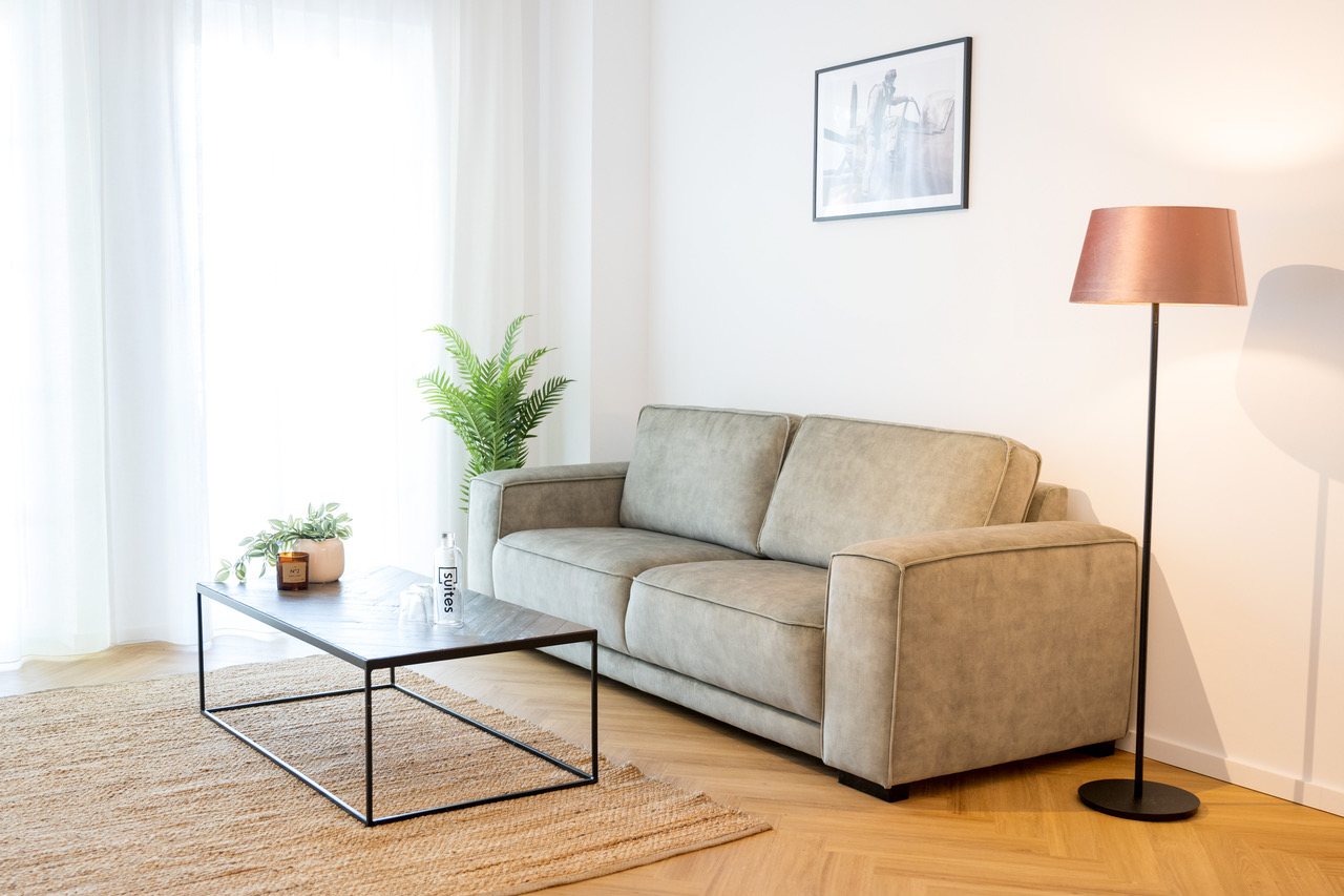 Wohnbereich mit Sofa, Stehleuchte und Couchtisch vor hellen Vorhängen